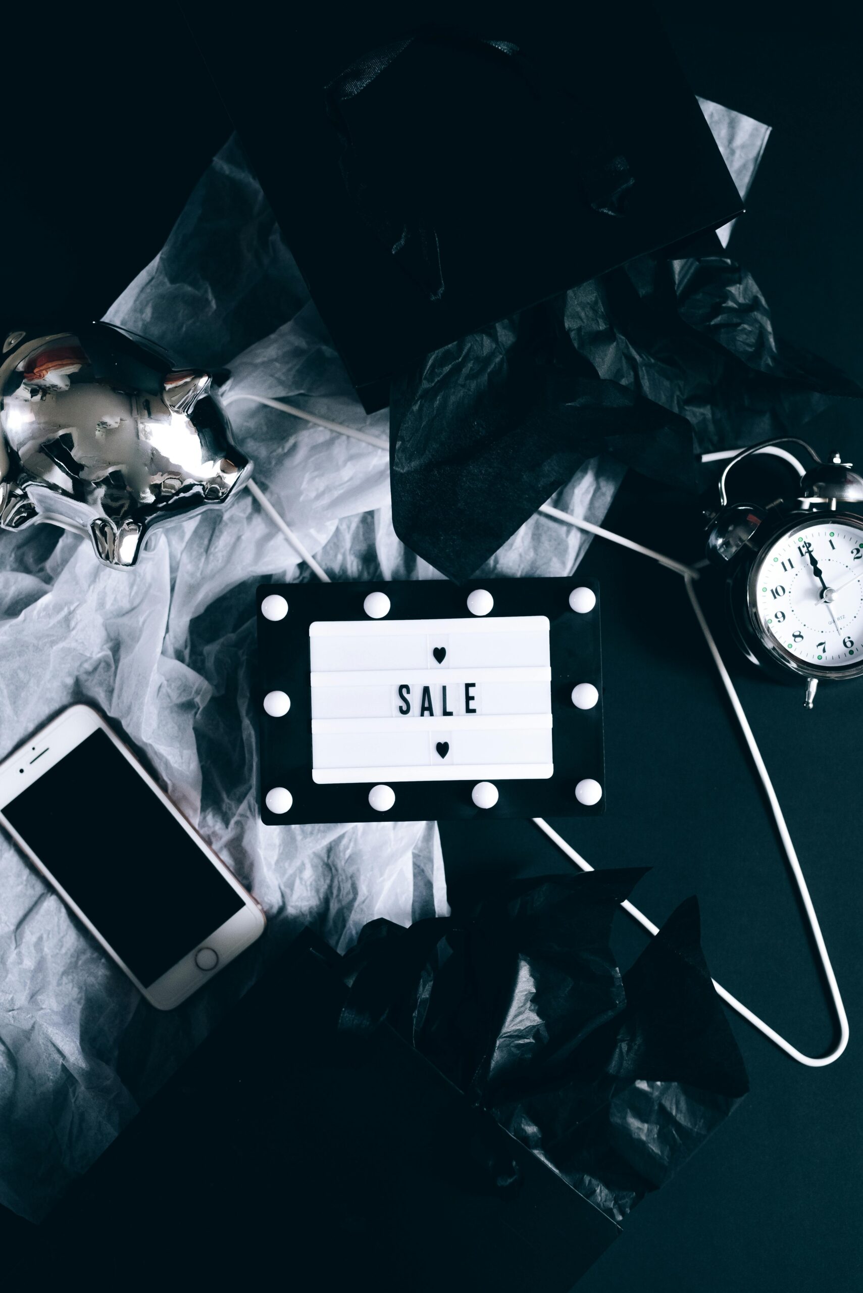 Stylish and dark flat lay featuring a sale sign, smartphone, retro alarm clock, and piggy bank.